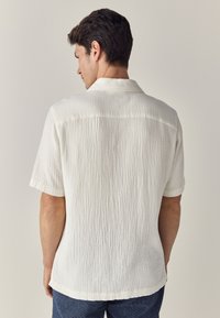 Camicia bianca a maniche corte con colletto e taglio dritto. I cuciture visibili e un leggero motivo stropicciato arricchiscono il design.