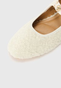 Chaussure à enfiler en crème texturée avec un bout arrondi, un intérieur beige, et une sangle décorative ornée de perles et d'accents dorés.