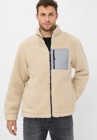 Giacca in pile beige con superficie testurizzata, dotata di tasca a zip grigia sul petto e zip nera. Maniche lunghe e design con colletto alto.