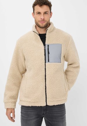 Beige fleecejakke med en tekstureret overflade, der har en grå lynlåslomme på brystet og sort lynlås. Lange ærmer og høj krave-design.
