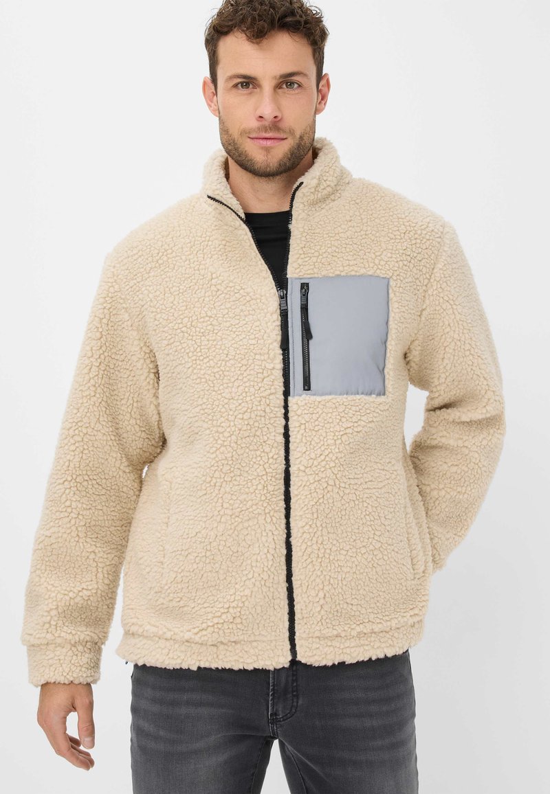 Giacca in pile beige con superficie testurizzata, dotata di tasca a zip grigia sul petto e zip nera. Maniche lunghe e design con colletto alto.