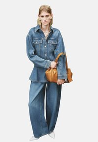 Giacca di denim con grandi tasche, chiusura a bottoni e maniche ampie, abbinata a pantaloni di denim larghi. Borsa marrone e scarpe bianche a punta.