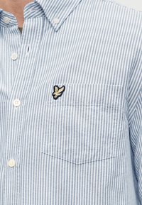 Ανοιχτό γαλάζιο και λευκό ριγέ πουκάμισο με κουμπιά, διαθέτει τσέπη στο στήθος και ένα μικρό κεντημένο λογότυπο σε ναυτικό μπλε και κίτρινο.