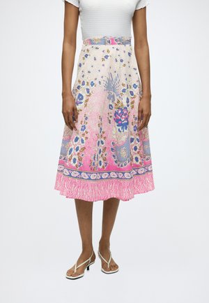 Femme portant une jupe fleurie mi-longue avec des motifs roses, bleus et blancs, associée à un haut blanc texturé à manches courtes et des sandales blanches.