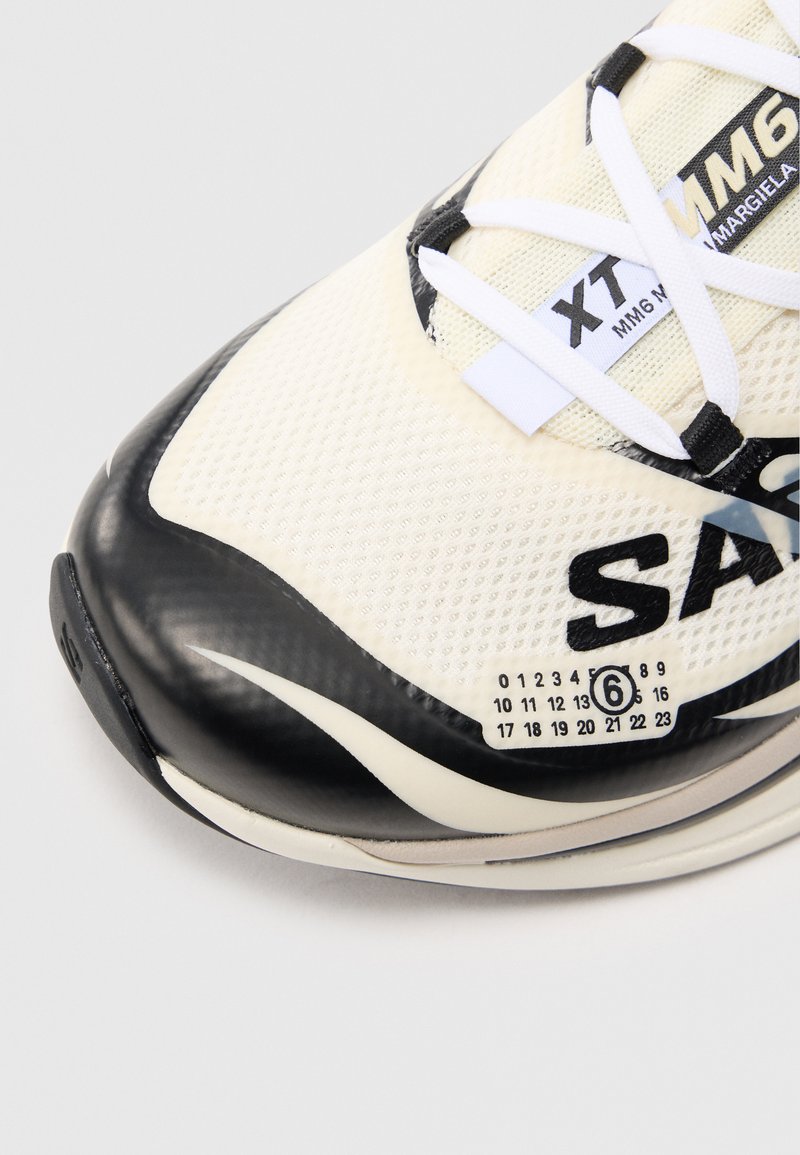 Hvide mesh sneakers med sorte gummidetaljer. Har snørebånd, en tekstureret overdel og en mønstret sål med numeriske detaljer i siden.