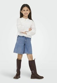 Witte blouse met lange mouwen en kantdetails, denim bermuda shorts en hoge bruine leren laarzen met ritsen. Staande met de armen gekruist.