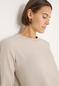 Beżowa bluzka z długim rękawem wykonana z teksturowanej tkaniny, z falbaną przy dekolcie. Charakteryzuje się prostym, luźnym krojem, bez widocznych wzorów czy ozdób.
