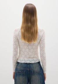 Top in pizzo a maniche lunghe di un beige chiaro con motivo floreale, abbinato a pantaloni in denim blu a vita alta con ampie tasche posteriori.