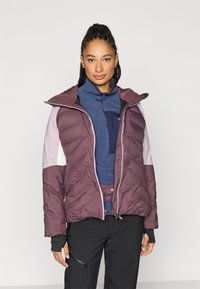 Podložena jakna v vijoličnih tonih z našito kapuco v kontrastni barvi, žepki z zadrgami in teksturiranimi paneli. Nošena preko modrega srednjega sloja.
