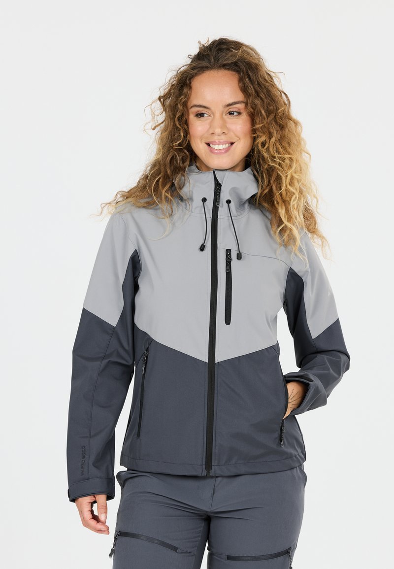 Femme aux cheveux bouclés portant une veste d'extérieur bicolore grise et un pantalon assorti, souriante et posant avec une main dans la poche.