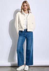 Witte fleece jas met hoge kraag en zilveren drukknoopjes, gecombineerd met losse blauwe spijkerbroeken en witte sneakers.