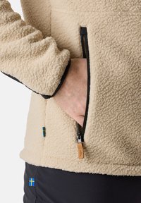 Beige fleece jacket with a zippered pocket and black accents. Features a soft texture and small blue Sweden flag detail on the sleeve.