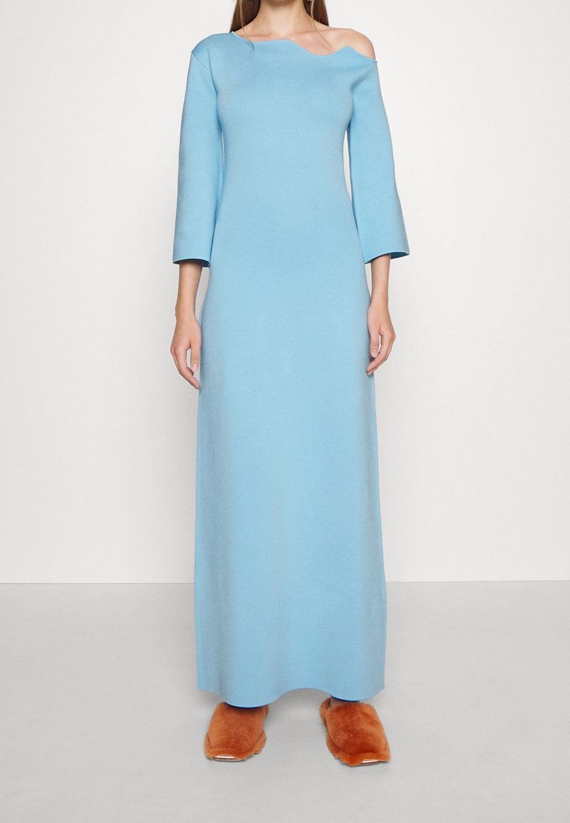 Femme portant une robe maxi bleu clair à manches longues et épaules dénudées ainsi que de grandes chaussons bruns en fourrure, debout devant un fond blanc uni.