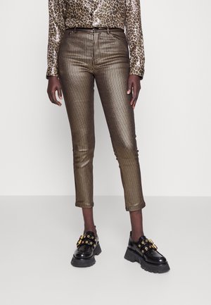 Persoon draagt metalen goud- en zwart gestreepte broek, luipaardprint shirt en zwarte chunky schoenen met gouden gespen, staand tegen een effen achtergrond.