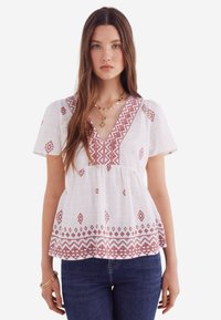 Witte blouse met korte mouwen en V-hals, versierd met rode geometrische borduursels. Gathered taille, lichtgewicht stof en een relaxte fit. Gecombineerd met blauwe spijkerbroek.