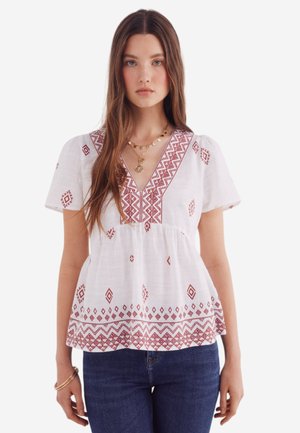 Blouse blanche à manches courtes avec décolleté en V et broderie géométrique rouge. Taille froncée, tissu léger et coupe décontractée. Associée à un jean bleu.