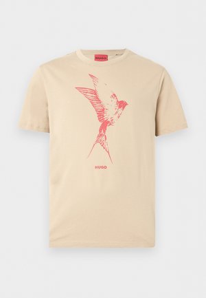 Beige T-skjorte med korte ermer med en rød fuglegrafikk på brystet og "HUGO" trykt under fuglen.