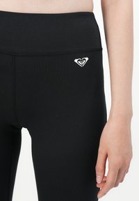 Svart tights laget av elastisk stoff med høy midje og en liten hvit logo på venstre side, med flate sømmer og jevn tekstur.