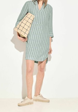 Kobieta w jasnopistacjowej sukience w paski, trzymająca beżową plecioną torbę, ubrana w jasnobeżowe trampki, stojąca na białym tle.