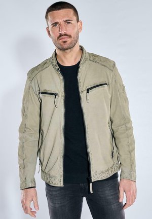 Adult man with short hair and beard wearing a beige zip-up jacket over a black shirt, standing against a plain light background.