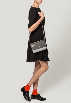 Vestido negro con mangas cortas, bolso de cadena plateada, acabado texturizado gris, calcetines naranjas, zapatos de charol, pose de pie contra un fondo claro.