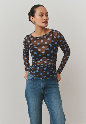 Femme portant un haut noir transparent à manches longues avec des pois bleus sur une couche noire, assorti à un jean bleu en denim, mains dans les poches.