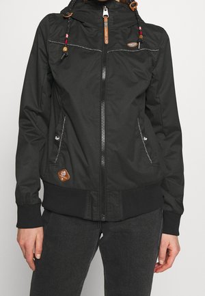 Sort lynlåsjakke med hætte og perlede snore, to forlommer, ribbede ærmeender og kant, båret over mørkegrå bukser.