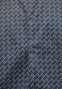 Navyblauw shirt met witte zigzagpatroon, voorzien van twee knopen en een gladde stoftextuur. Het ontwerp bedekt het gehele oppervlak uniform.