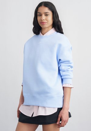 Naine pikkade tumedate juustega, kes kannab hele sinist dressipluusi kahvatu roosa särgi ja musta seeliku üle, seistes neutraalse tausta ees.