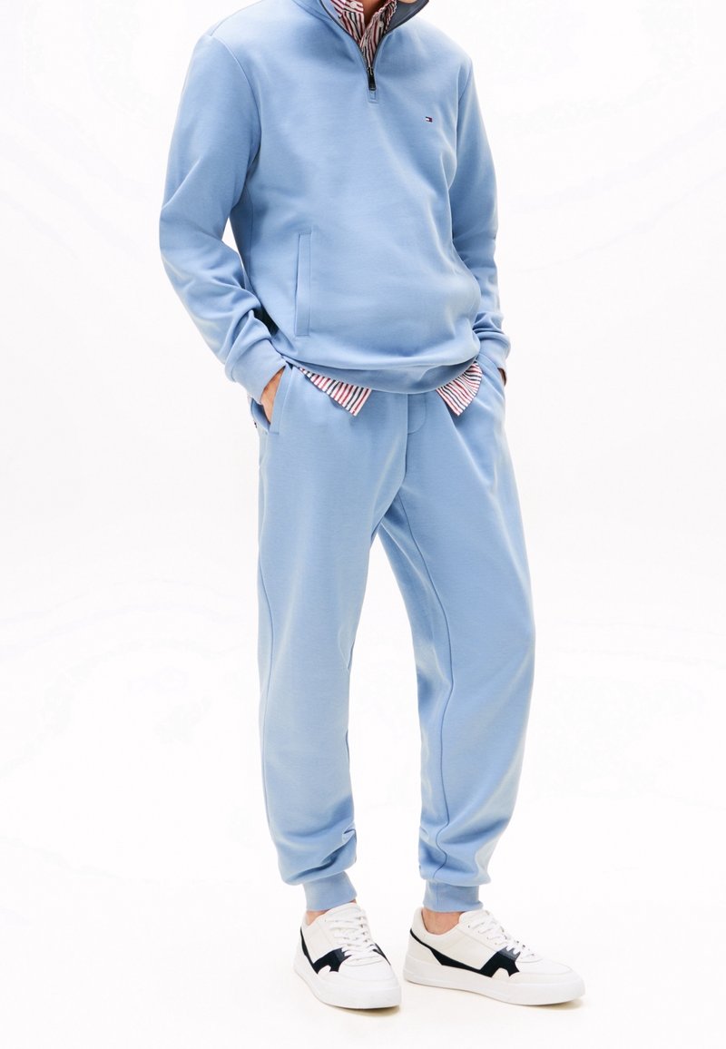 Personne portant un sweat-shirt bleu clair à fermeture éclair quart de zip et un pantalon de jogging assorti avec des baskets blanches et une chemise rayée rouge et blanche partiellement visible.