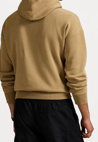 Person iført en kamelfarvet hættetrøje med lange ærmer og sorte bukser med en baglomme med knap, set bagfra.