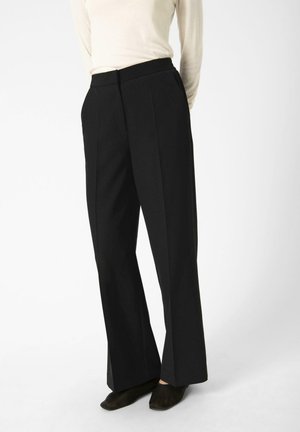 Pantaloni neri a gamba larga con una texture liscia, caratterizzati da un'alta vita e pieghe frontali. Abbinati a scarpe scure e flat.