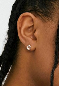 Pendiente de plata con un diseño circular entrelazado. Primer plano de un peinado trenzado oscuro, mostrando el pendiente sobre un tono de piel marrón.