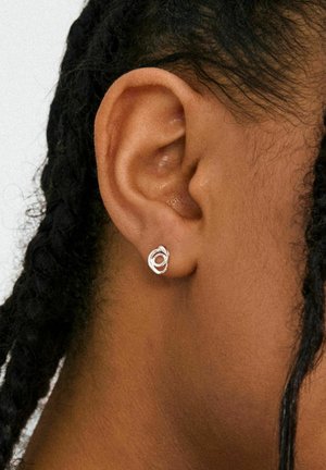 Zilveren steekoorbel met een verweven cirkelontwerp. Close-up van een donkere gevlochten haarstijl, waarbij de oorbel goed zichtbaar is op een donkere huidtint.