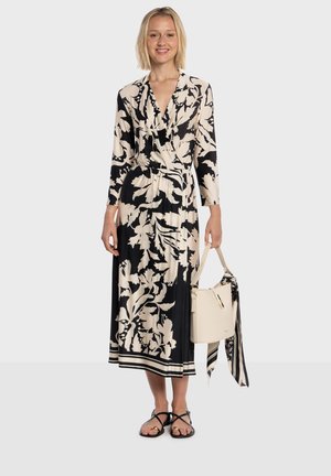 Robe à manches longues avec imprimé floral crème et noir, taille cintrée, longueur jusqu'aux chevilles, associée à un sac à main blanc et des sandales noires à lanières.