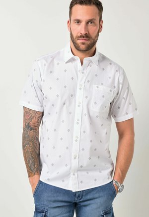 Uomo che indossa una camicia bianca a maniche corte con un piccolo motivo a rombi e pantaloni cargo blu con le mani nelle tasche.