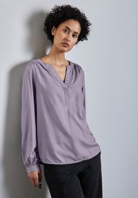 Ljuslila blus med lång ärm, V-ringning och gatherade detaljer vid axlarna. Buren med mörka jeans mot en neutral vägg.