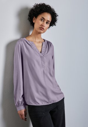Blusa de manga larga en púrpura claro con cuello en V y detalles fruncidos en los hombros. Llevada con jeans oscuros frente a una pared neutral.