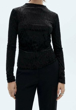 Mujer con una camiseta ajustada de manga larga negra con un sutil estampado de leopardo, combinada con pantalones negros, con las manos relajadas a los lados.