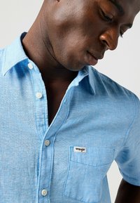 Camicia azzurro chiaro a maniche corte in lino con colletto button-down, tasca frontale e un piccolo logo "Wrangler" sulla tasca.