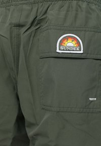 Bañadores verdes hechos de material ligero, cuentan con una cintura elástica y un bolsillo trasero con un colorido parche del logo "Sundek".