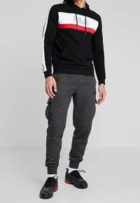 Sudadera negra con rayas horizontales blancas y rojas, con el logo de "FILA". Pantalones deportivos grises con bolsillos laterales, combinados con zapatillas deportivas negras.