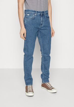 Mężczyzna ubrany w niebieskie, proste dżinsy i brązowe tenisówki na sznurowadła, stojący na tle jednolitego białego tła.