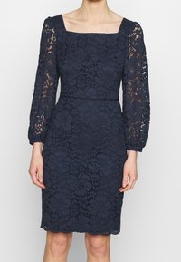Navy lace dress with long puffed sleeves and a square neckline. Fitted silhouette with a scalloped hem and floral patterns throughout.