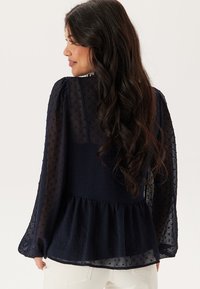 Blusa peplum blu navy con texture a pois, maniche lunghe trasparenti e orlo arricciato. Presenta un collo rotondo e un corpino aderente.