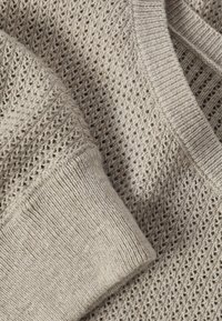 Béžová pletená látka svetru s žebrovaným lemem a výstřihem do V, zdobená texturovaným vzorem s otevřeným propletením.