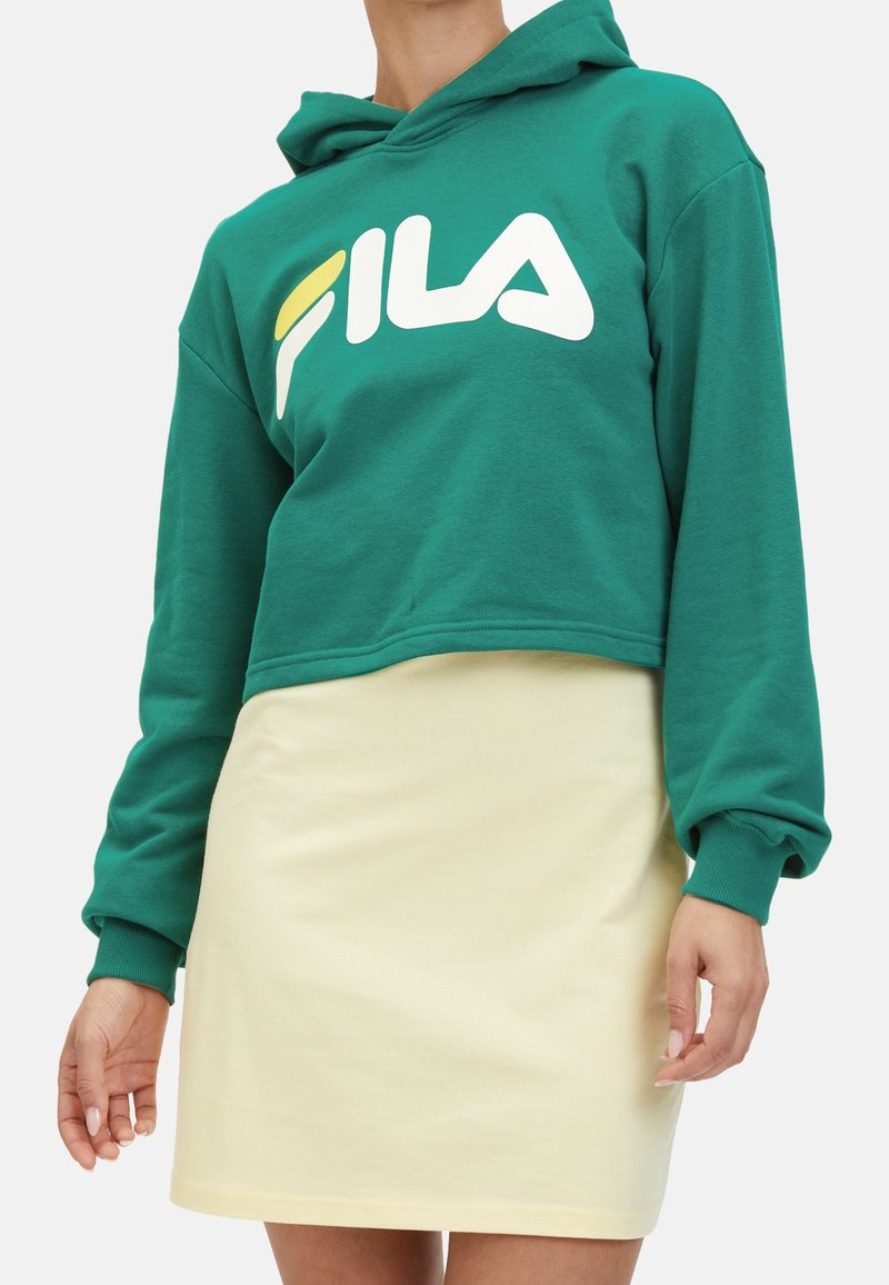 Modella che indossa un hoodie cropped verde con un logo "FILA" bianco e giallo, abbinato a una mini gonna gialla chiara.