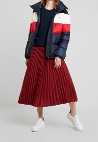 Vadderad marinblå jacka med röda och vita horisontella ränder, ihopparad med en röd plisserad midikjol och vita sneakers med gröna accenter.