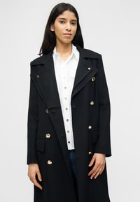 Νέα γυναίκα που φοράει μαύρο διπλόμπουτο παλτό με χρυσά κουμπιά πάνω από λευκό πουκάμισο και μπλε τζιν, μπροστά σε απλό φόντο.