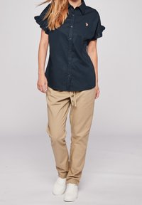 Mörkblå kortärmad skjorta med volangärmar och en liten broderad detalj, ihopparad med beige byxor med dragsko och vita sneakers.